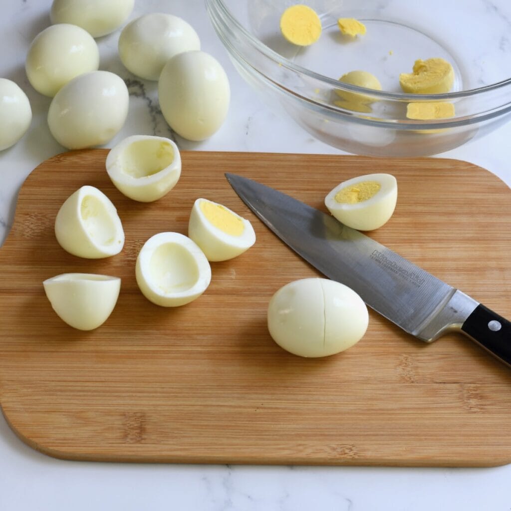 Hand slicing a hard-boiled egg crosswise on a wooden cutting board with yolks in a bowl nearby