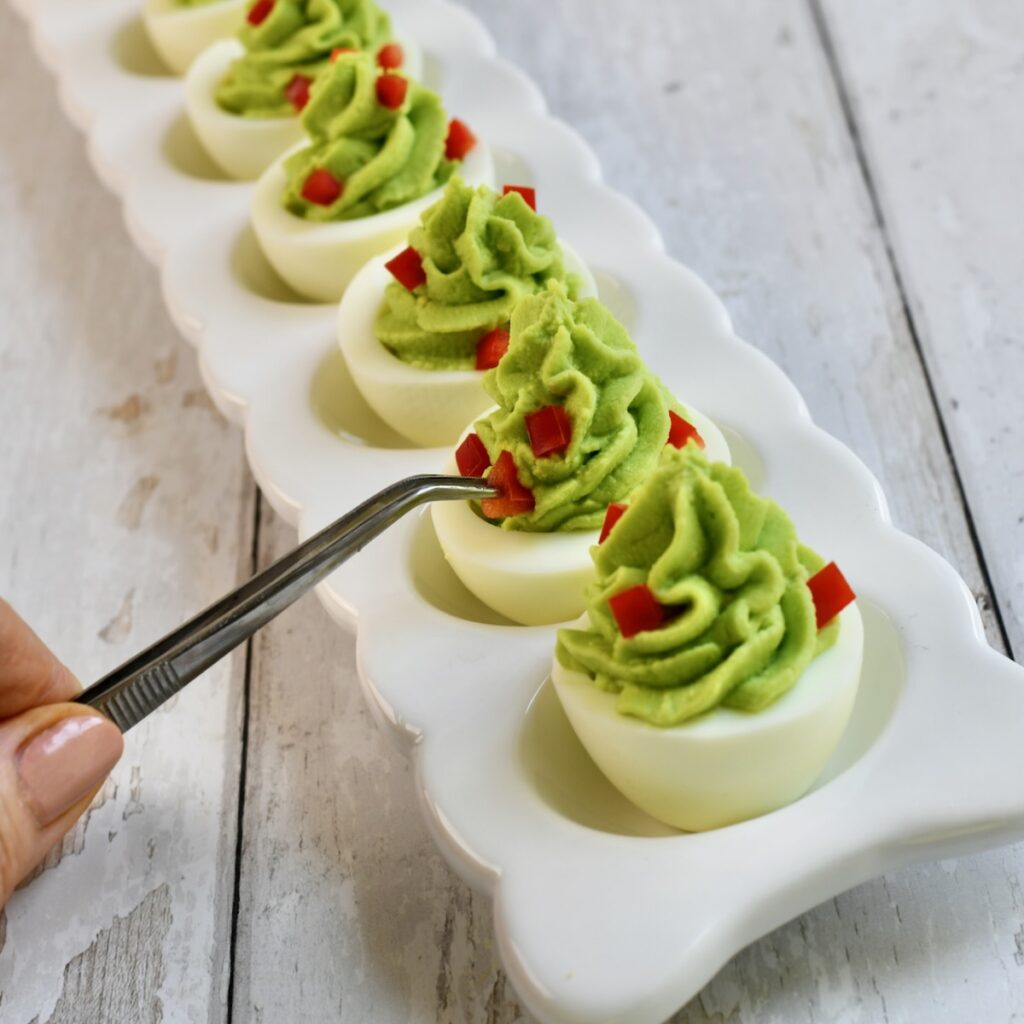 hand using tweezers to place tiny red pepper pieces as ornaments on piped christmas tree deviled eggs