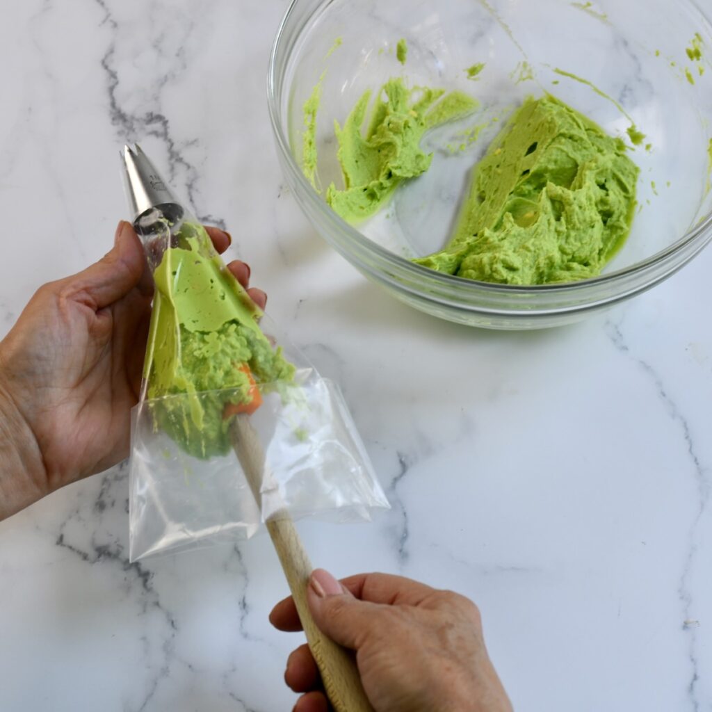 Hand using a spatula to transfer green avocado-yolk mixture into a pastry bag with a piping tip