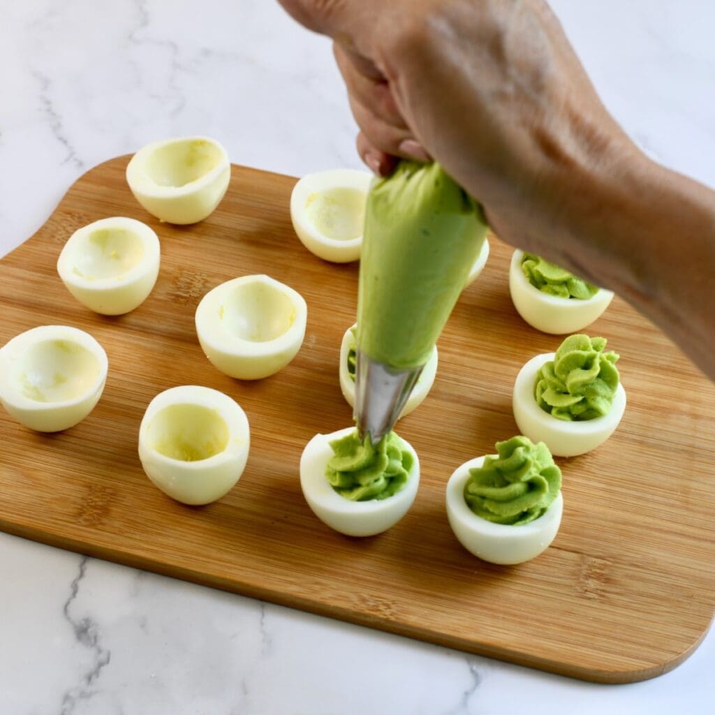 Hand piping avocado and yolk filling into egg white halves on a wooden cutting board
