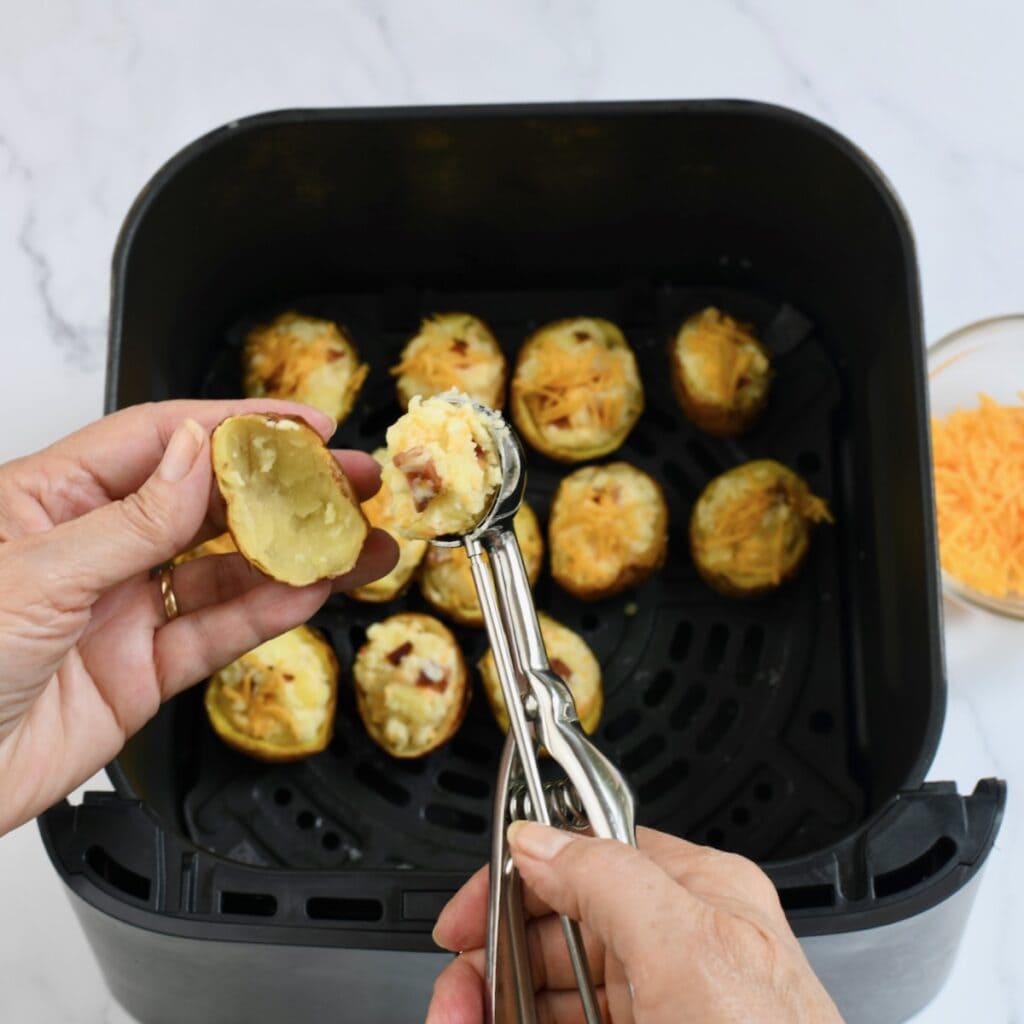 Hands using a cookie scoop to fill hollowed potato skins with cheesy mashed potato mixture inside an air fryer basket 