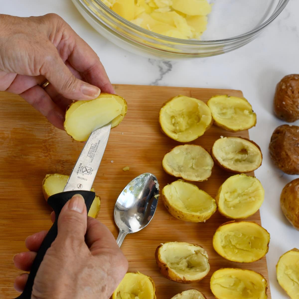 Hands using a paring knife to cut and scoop the center from cooked mini potatoes on a wooden board