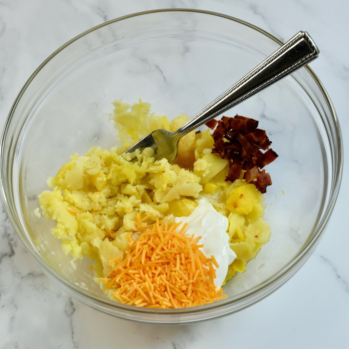 Shredded Cheddar cheese, sour cream, bacon, and mashed potatoes in a glass bowl with a fork, ready to mix for the potato filling