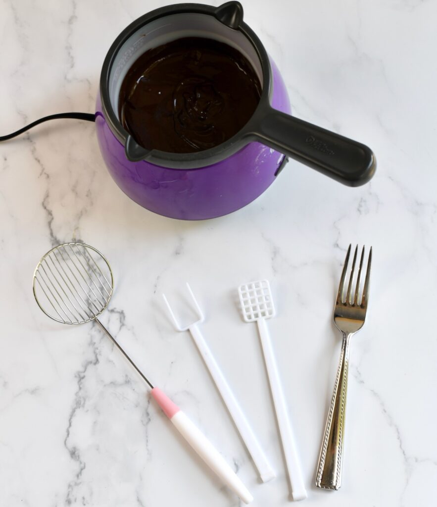 Melted chocolate in a purple melting pot with candy dipping tools and a fork placed on the counter