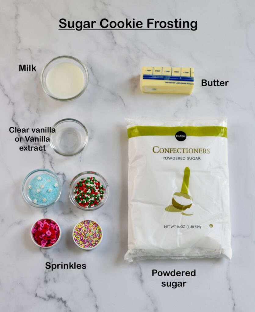 ingredients for sugar cookie frosting arranged on a marble surface, including milk, butter, powdered sugar, clear vanilla extract, and colorful sprinkles