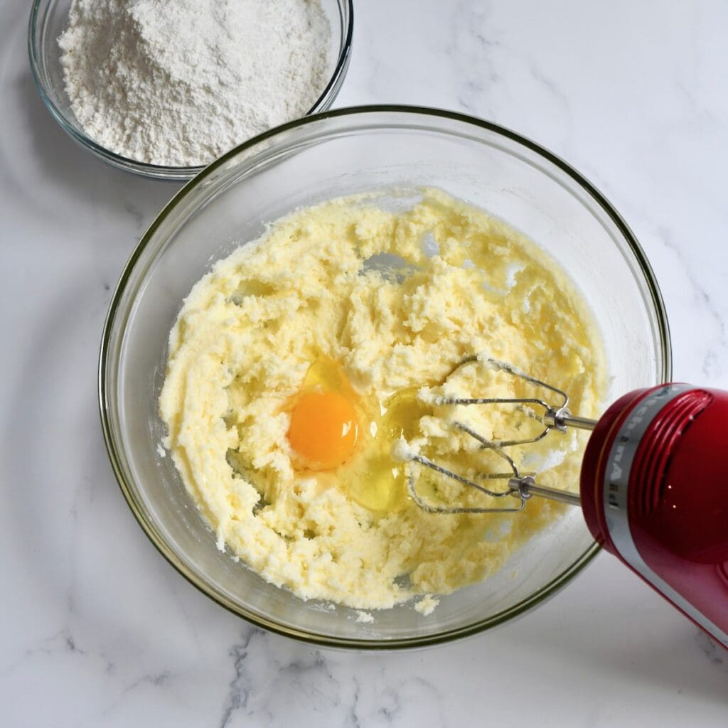 mixing bowl with creamed butter and sugar, an egg on top, and a hand mixer with flour in a separate bowl nearby