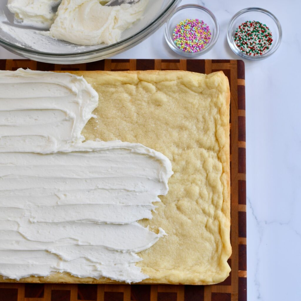 spreading white frosting over baked sugar cookie bars with bowls of sprinkles nearby