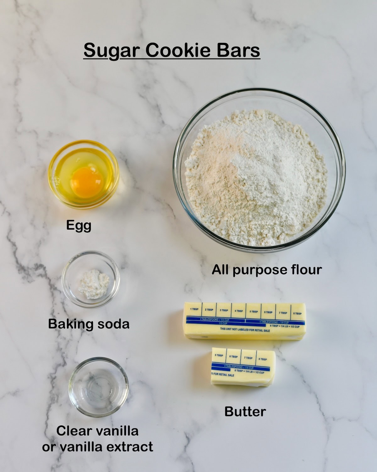 ingredients for sugar cookie bars displayed on a marble surface, including flour, an egg, baking powder, vanilla extract, and sticks of butter