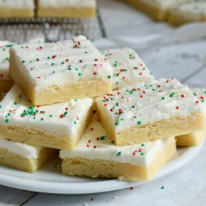 frosted sugar cookie bar squares with red and green sprinkles stacked on a white plate