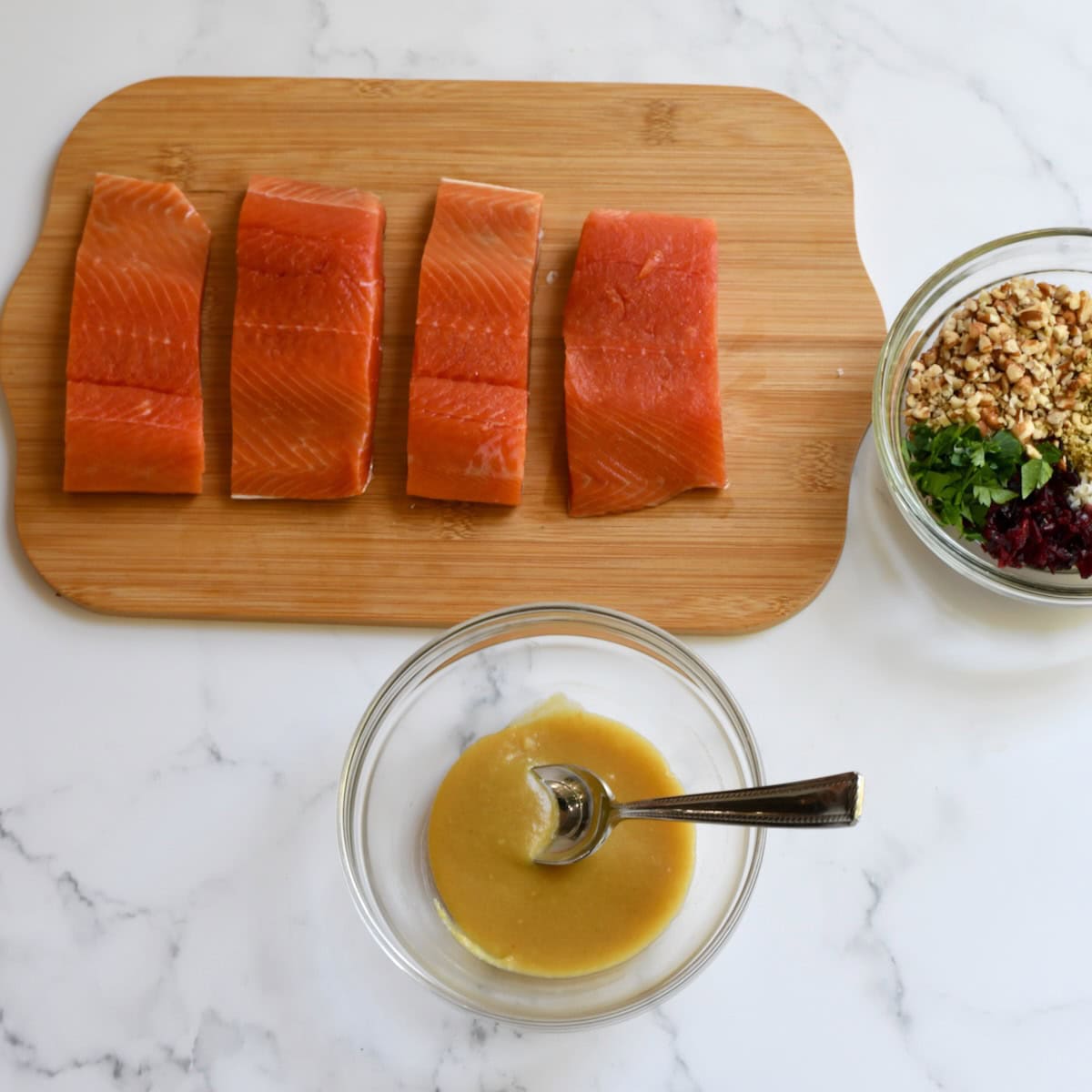 Four raw salmon fillets on a wooden cutting board with a small bowl of honey-Dijon mixture and another bowl containing chopped pecans, cranberries, Parmesan, and parsley