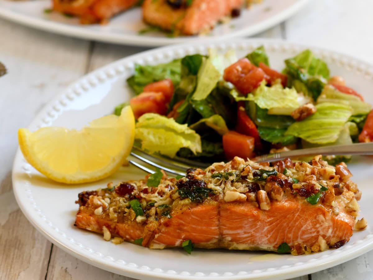 Pecan-crusted salmon served on a white plate with a green salad, diced tomatoes and a lemon wedge