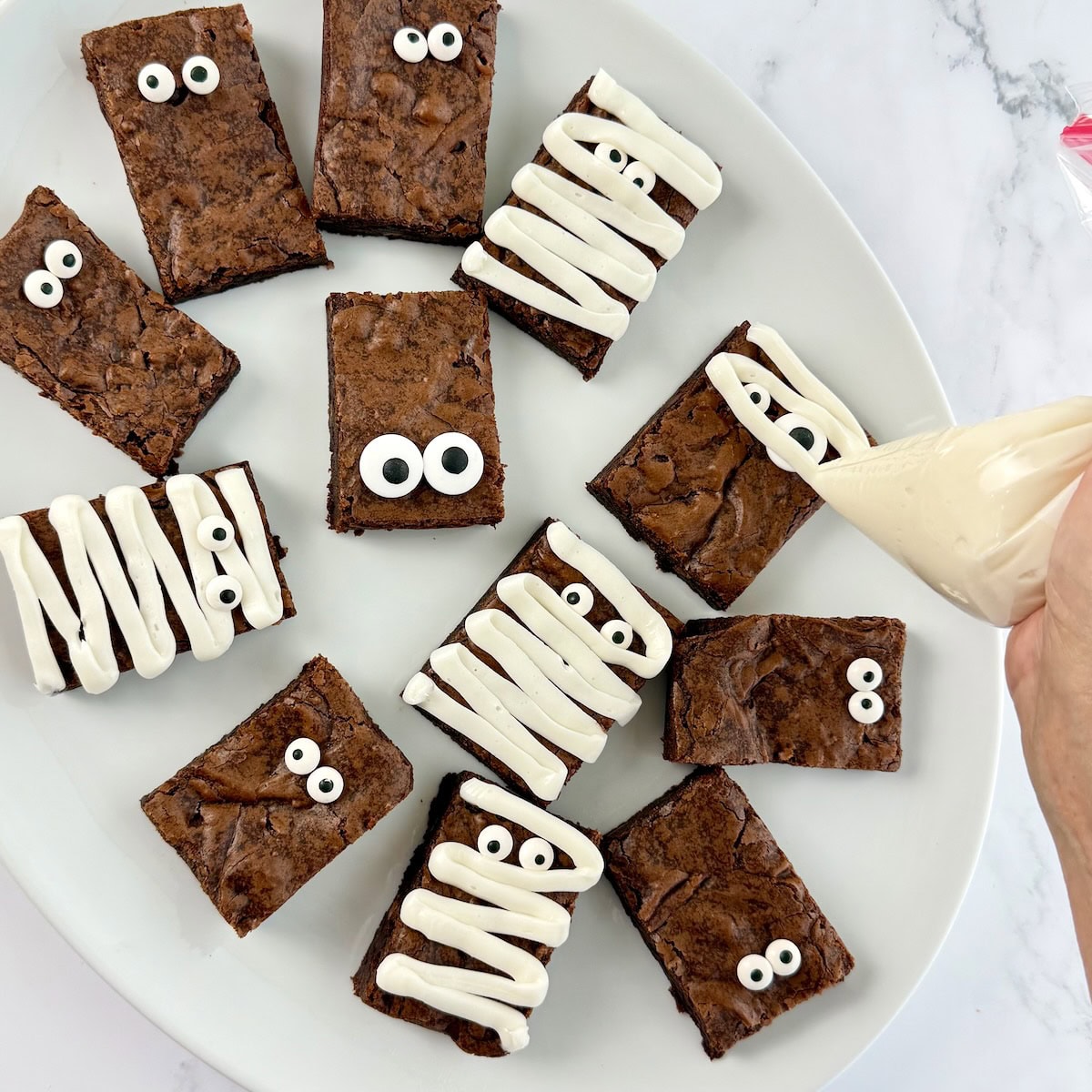 Pastry bag piping white frosting onto brownies with candy eyes.