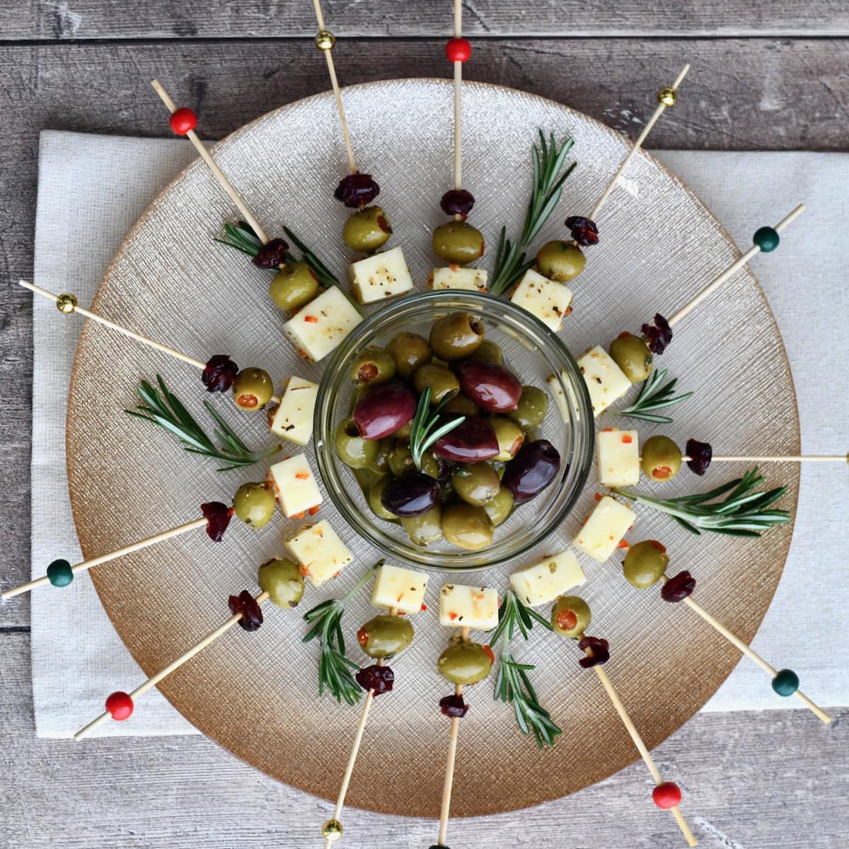 Overhead view of Cranberry Olive Cheese Bites arranged in a circular pattern around a bowl of mixed olives, garnished with rosemary sprigs on a gold platter