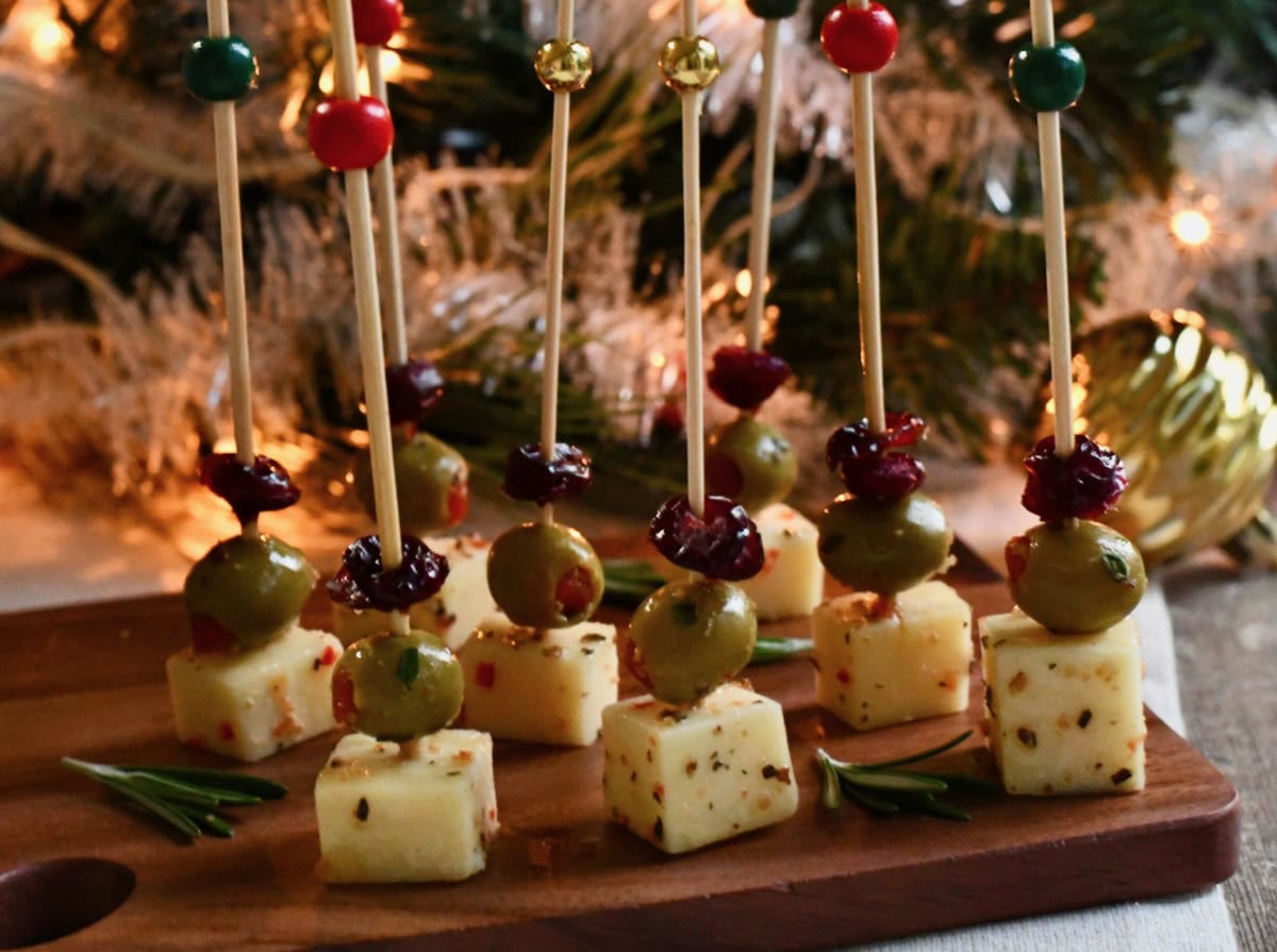 Cranberry Olive Cheese Bites arranged on a wooden serving board with rosemary sprigs, set in front of a Christmas tree decorated with lights and ornaments