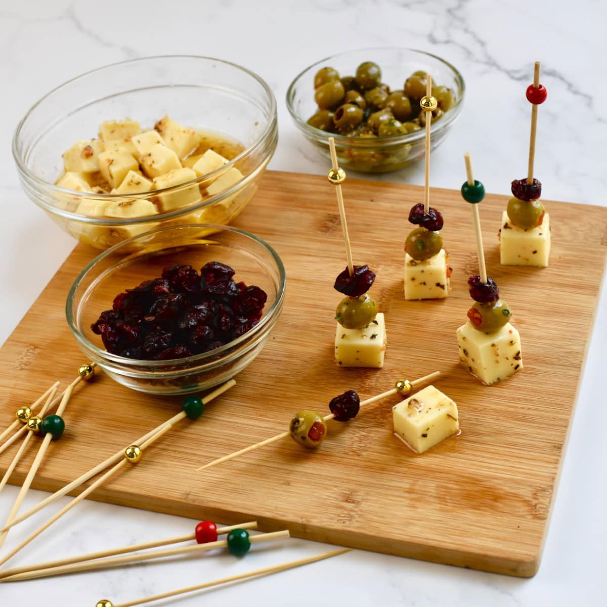Ingredients and partially assembled Cranberry Olive Cheese Bites on a wooden board with small bowls of cheese cubes, green olives, and dried cranberries
