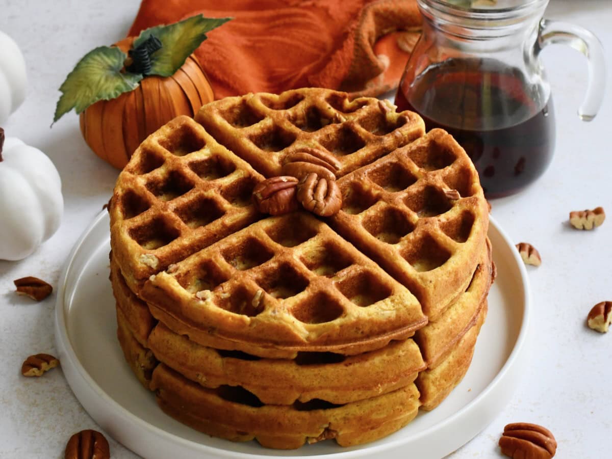 Stack of pecan pumpkin Belgian waffles on white plate with carafe of maple syrup in background.