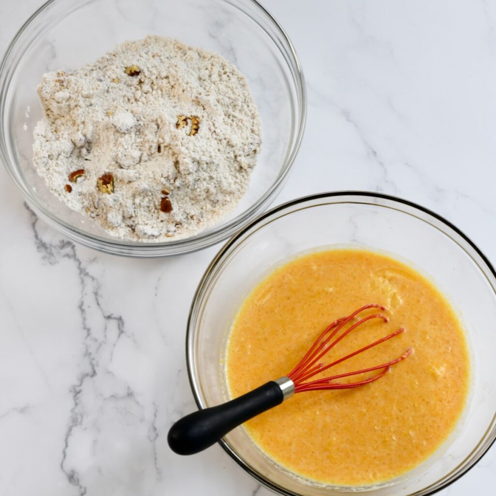 Whisk in a glass bowl of eggs and butter next to a glass bowl of waffle mix and nuts.