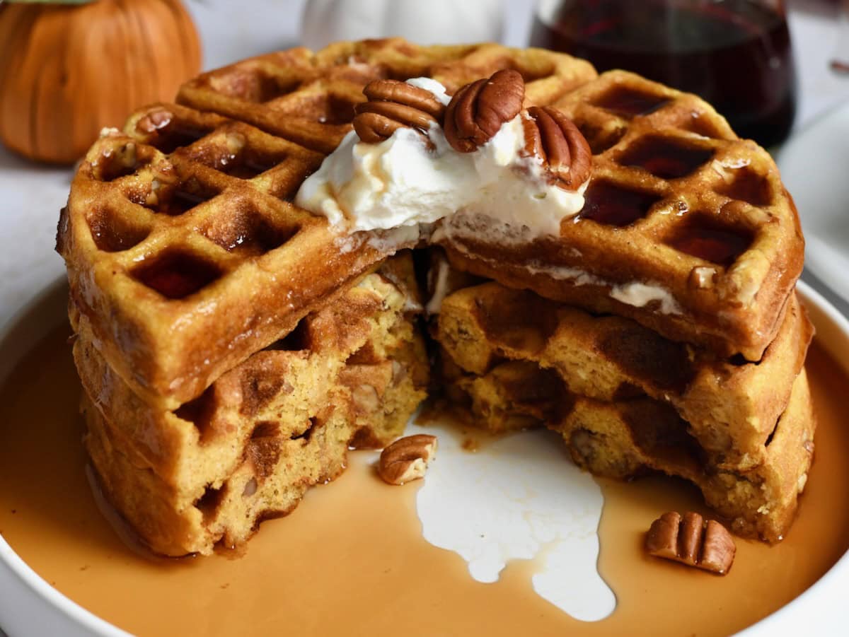 Stack of pumpkin pecan waffles sliced into and showing maple syrup and pecans on plate.