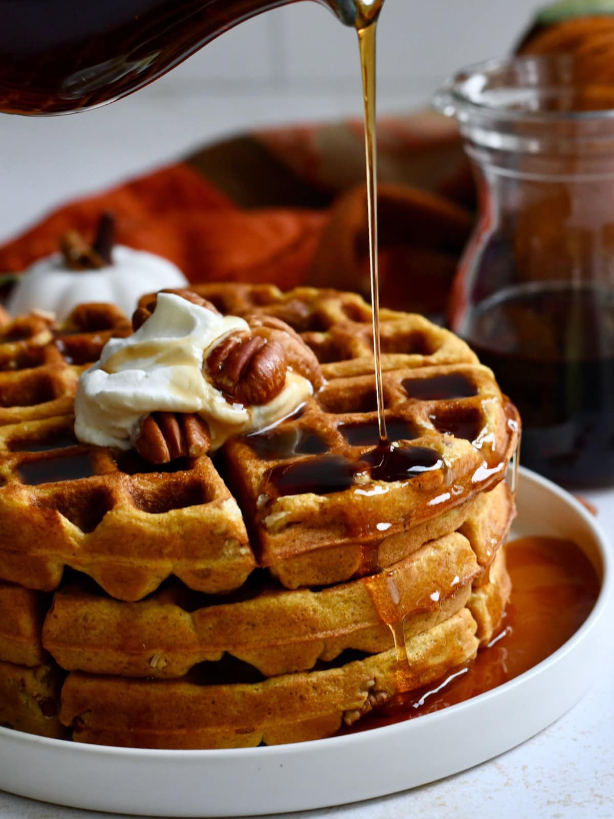 Maple syrup being poured over pumpkin pecan waffles with whipped cream and pecans.