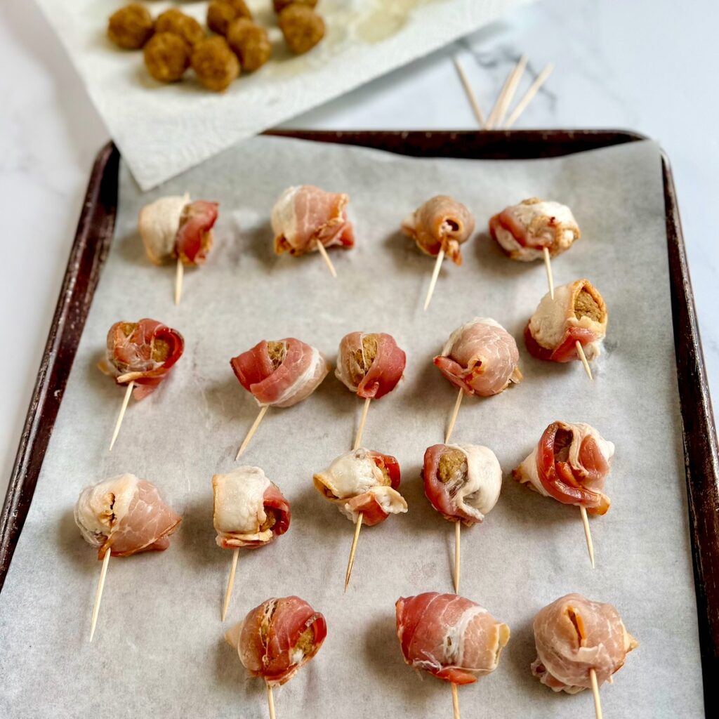 Meatballs wrapped in raw bacon and stuck with a toothpick and placed on a sheet pan.