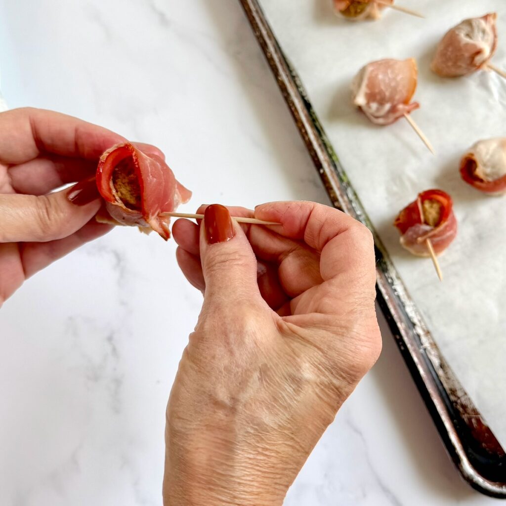 Hands sticking toothpicks into the meatball to keep the bacon wrapped around it.