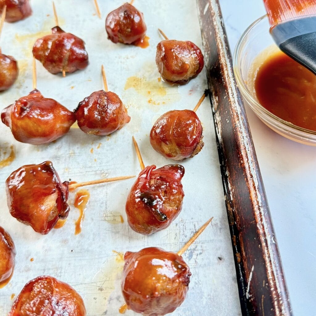 Brushing BBQ sauce onto the bacon wrapped meatballs on a sheet pan.