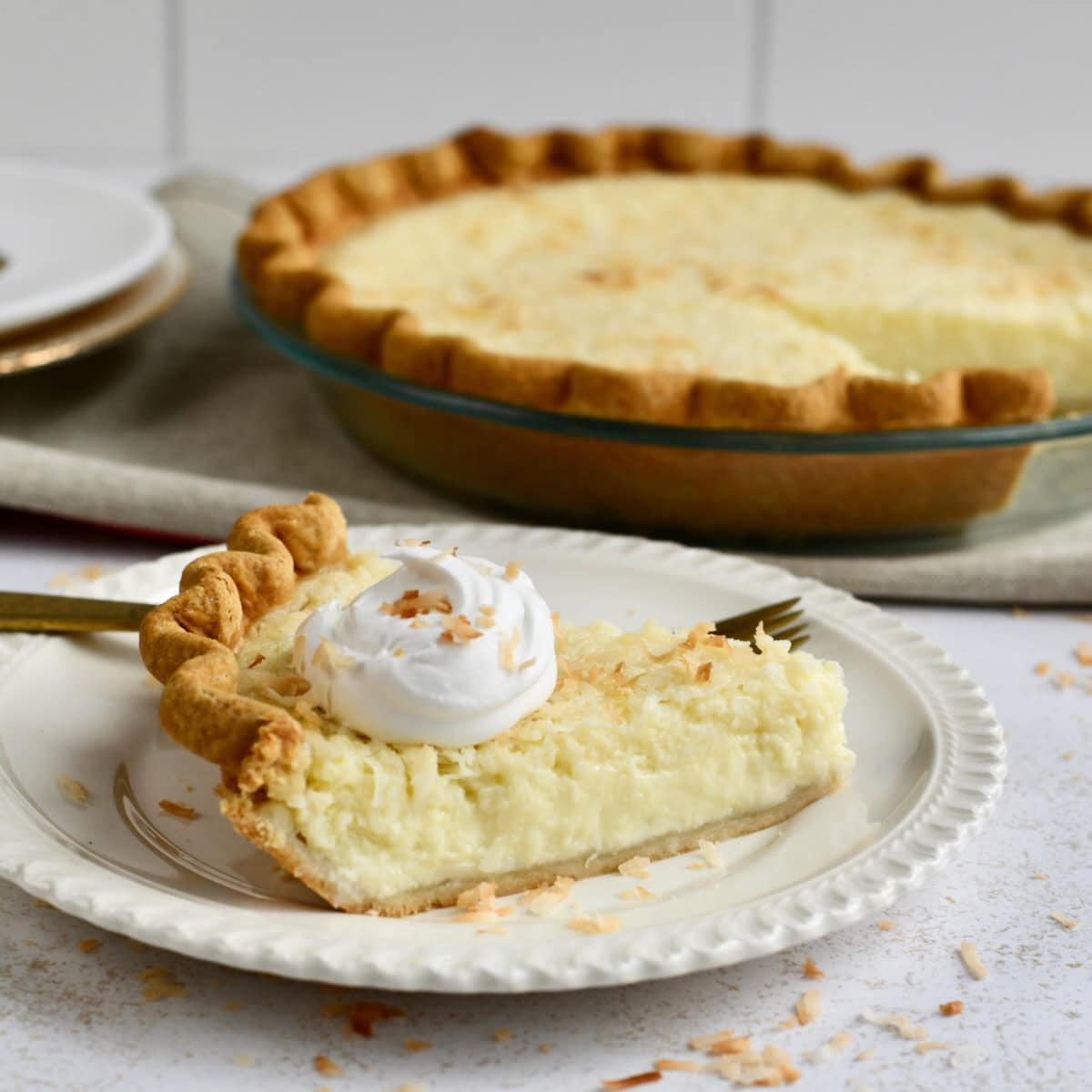 A slice of coconut custard pie with whipped cream on a white plate with the whole pie in the background.