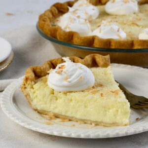 Close up image of a slice of coconut custard pie on a white plate with whipped cream on top. The whole pie is in the background.