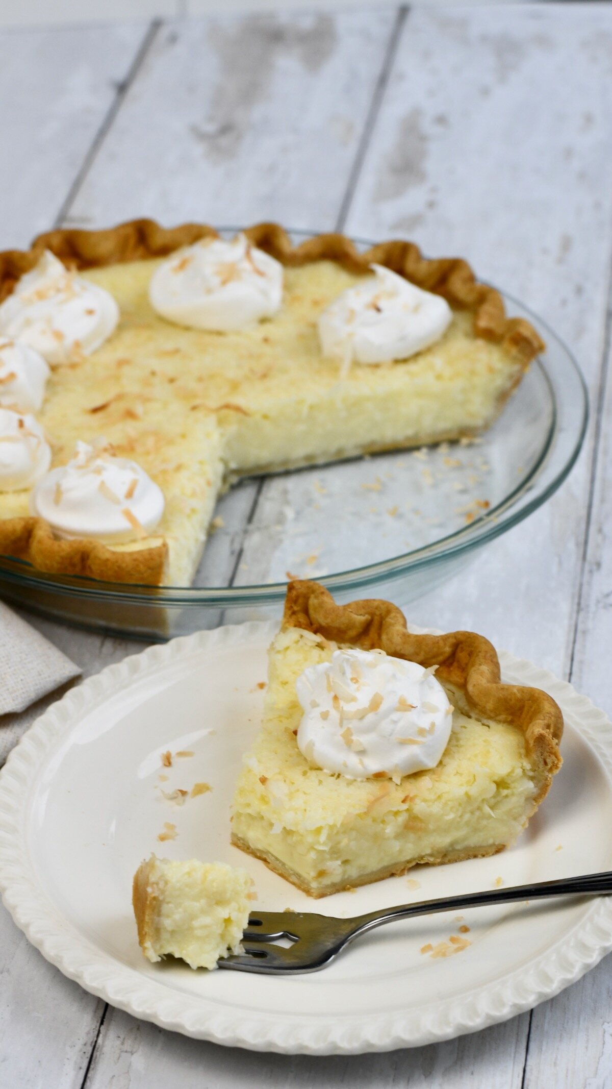 A slice of coconut custard pie with a fork taking a bite out on a white plate. The whole pie is in the background. 