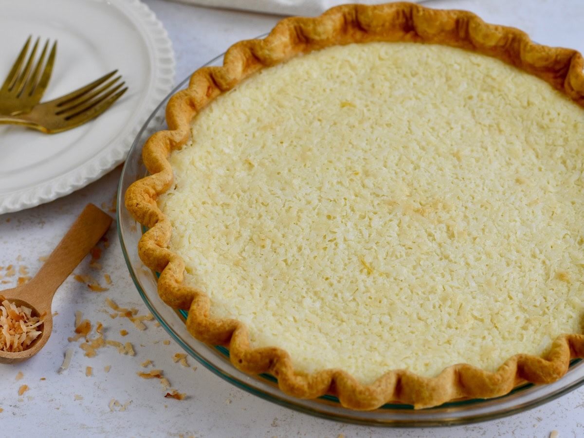 Close up image of coconut custard pie with white plate and gold forks in the background and a small wooden spoon on the side.