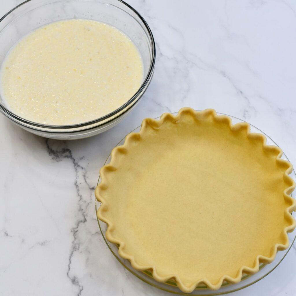 A unbaked pie crust crimped in a glass pie plate with a glass bowl of the coconut custard filling.