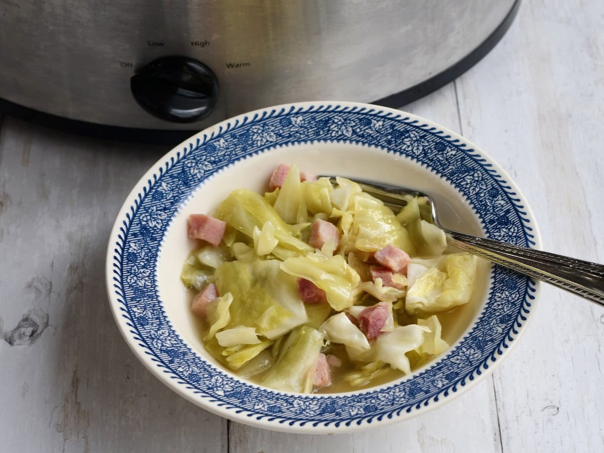 A white bowl with blue rim with crock pot ham and cabbage. A fork is resting on the side and the slow cooker is in the background.