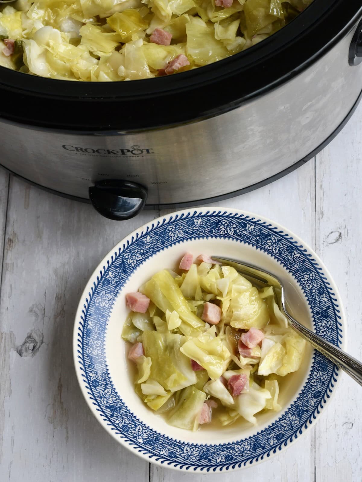A blue and white bowl of slow cooker cabbage and ham with a fork on the side and the slow cooker in the background.