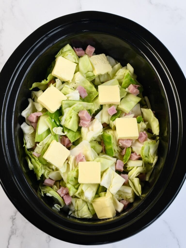 A black slow cooker bowl with cabbage, ham, and sliced butter.