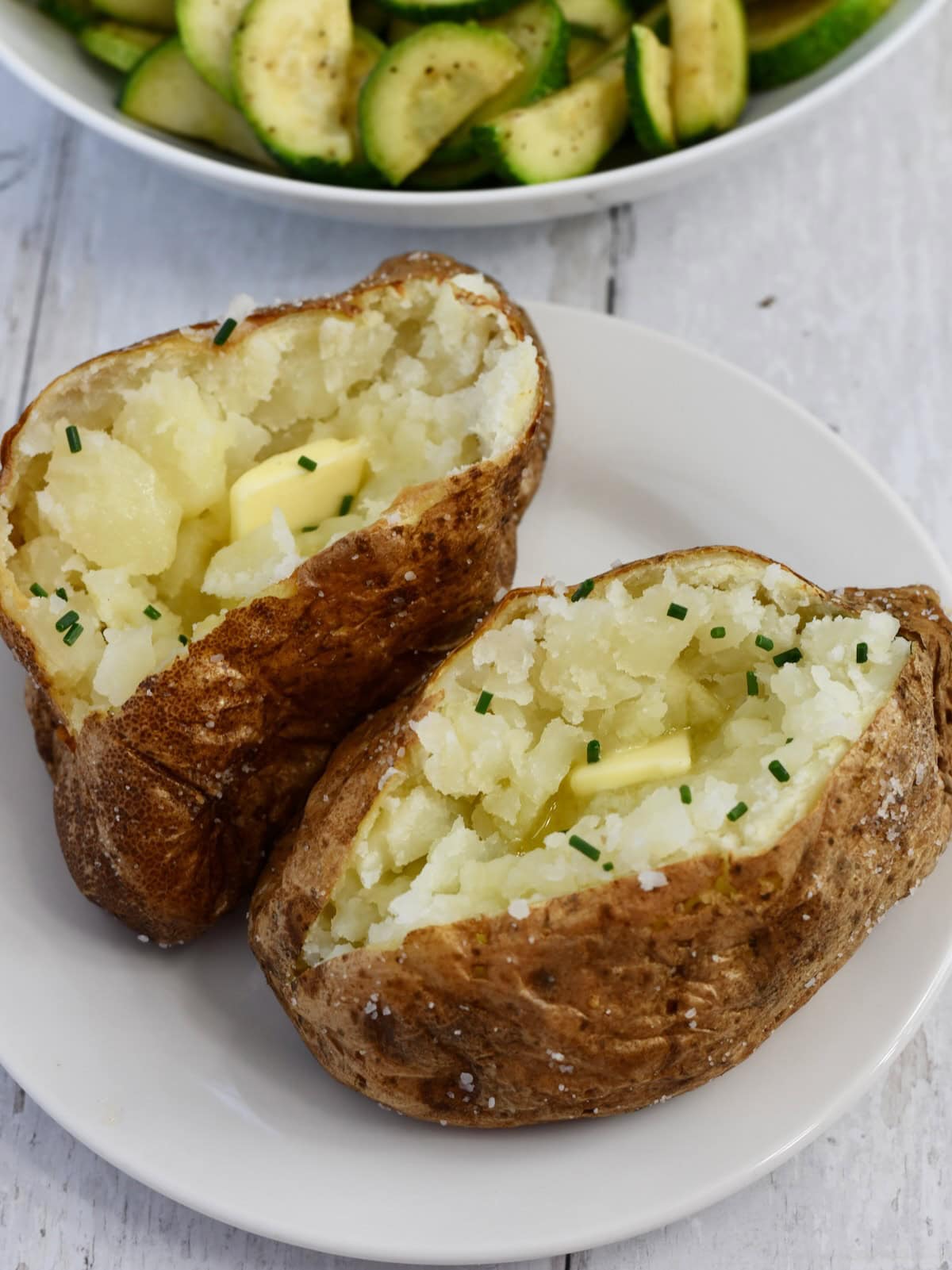 Two baked potatoes served on a plate both topped with butter and chives.