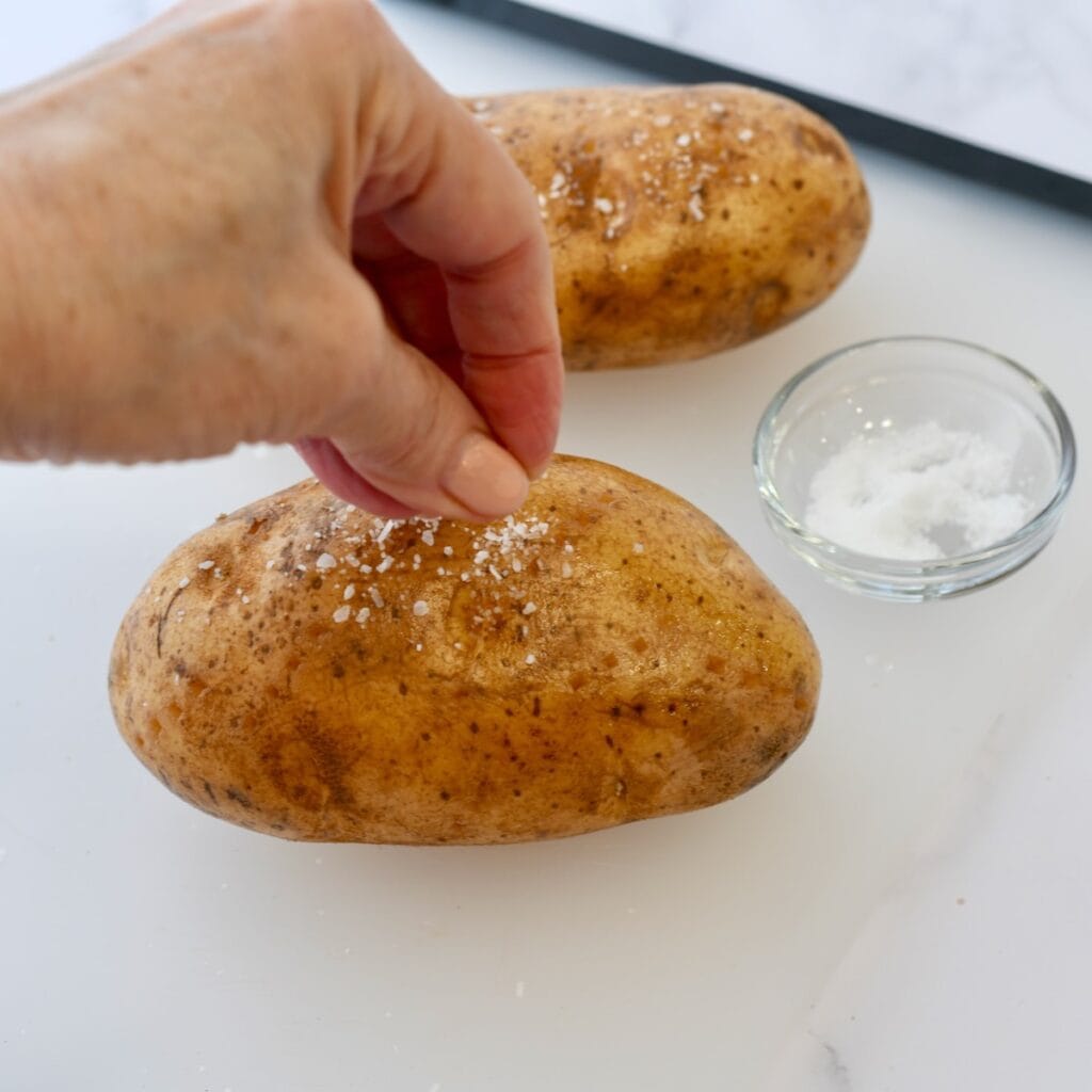 Sprinkling kosher salt on top of a russet potato on a cutting board.