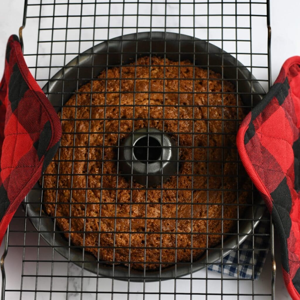 A baked carrot cake bundt cake in the bundt pan with wire cooling rack on top and plaid oven mitts.