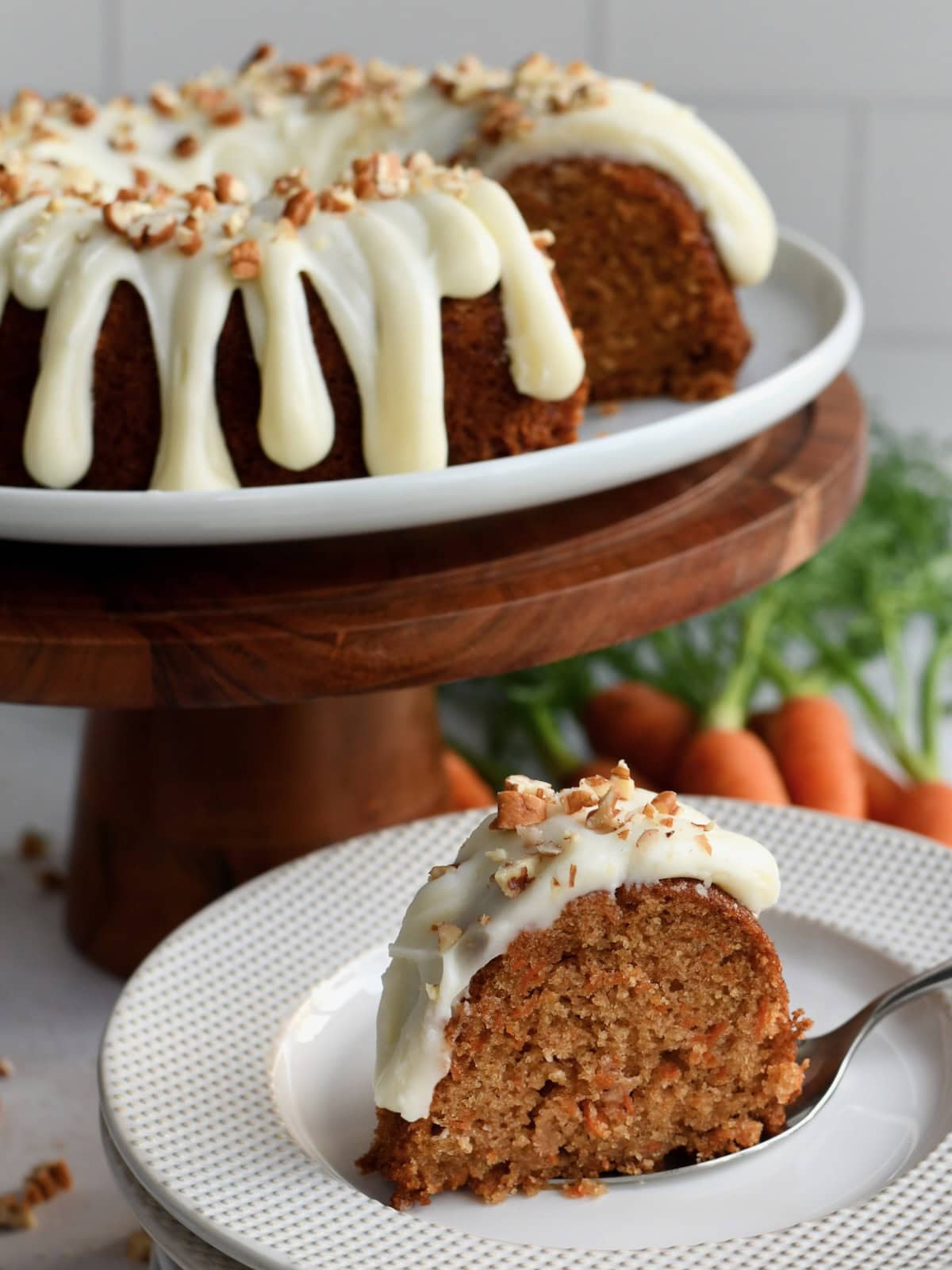 A carrot bundt cake with a slice of cake on a white plate with the cake on a cake stand in the backgrounbd.