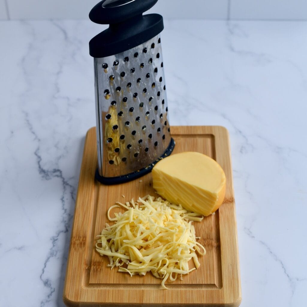 A cheese grater with gouda cheese being shredded on a wooden board.