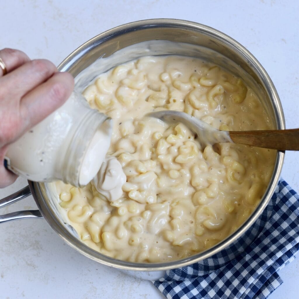A saucepan of gouda mac and cheese with a hand pouring alfredo sauce into the pot.