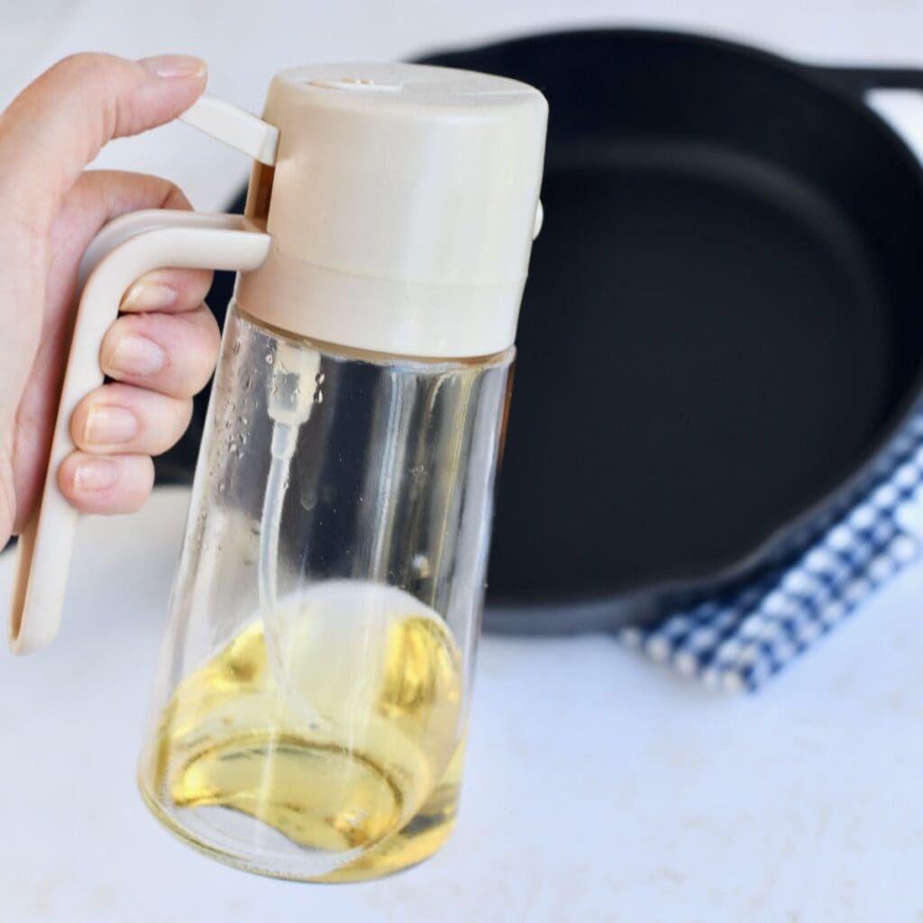 A hand with an oil spritzer with a cast iron skillet in the background on a blue plaid linen.