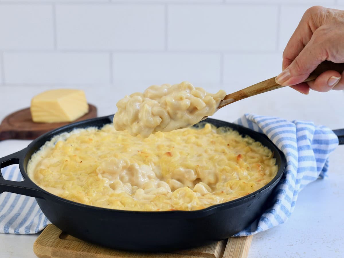 A cast iron skillet with gouda mac and cheese being scooped with a wooden spoon by a hand.