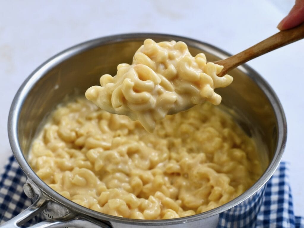 A metal saucepan with gouda mac and cheese being scooped on a wooden spoon. The pot is on a blue checkered linen.