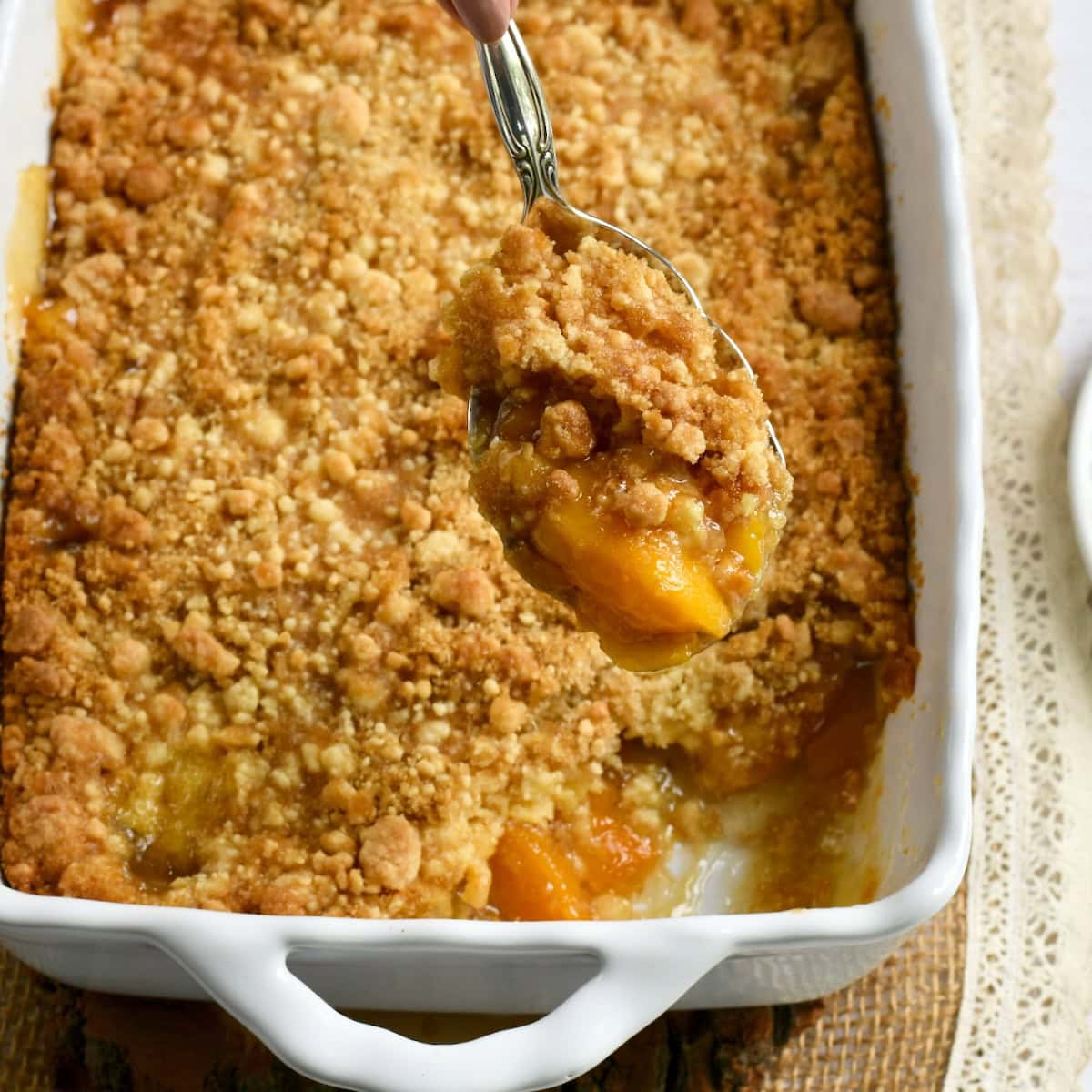 A spoon with cake mix peach cobbler with a white baking dish of the whole peach cobbler in the background.