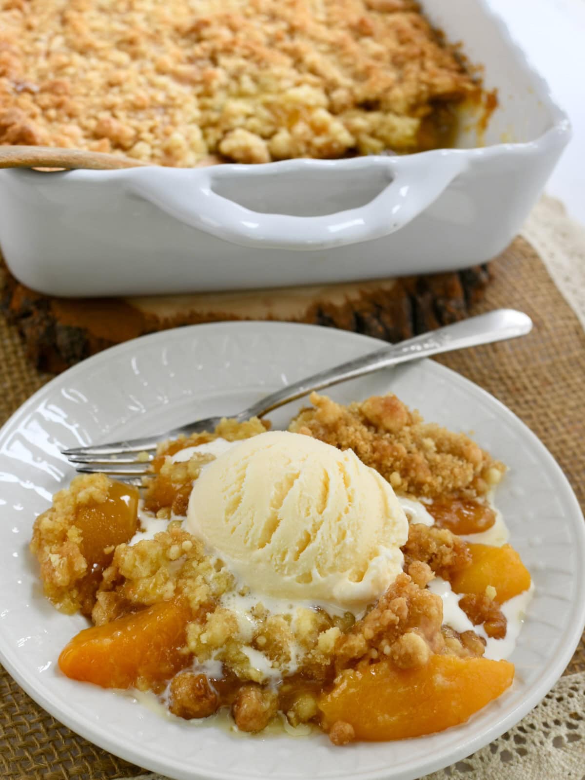 A white plate and fork with cake mix peach cobbler with a scoop of ice cream on top. The white baking dish of peach cobbler is in the background.