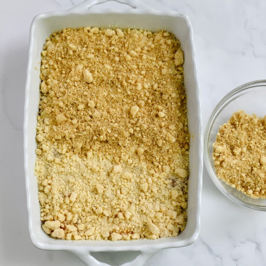 A white baking dish with the cake mix topping sprinkled on top of peach pie filling. A glass bowl of the extra cake mix mixture is to the side.