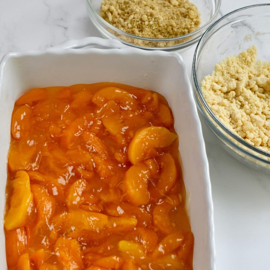 A white baking dish with peach pie filling. Two glass bowls of cake mix topping are to the side.