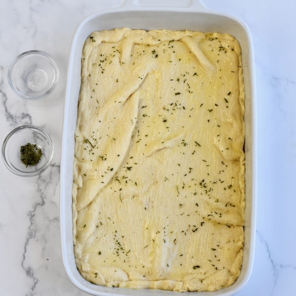 Layering crescent roll dough on top of the ham and cheese bake and brushing butter and herbs on top.