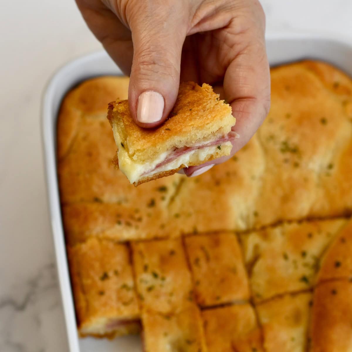 Hand taking a crescent roll ham and cheese square from the baking dish.