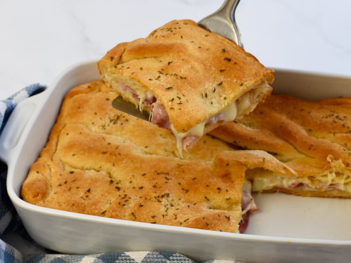 Crescent roll ham and cheese square being taken from the baking dish by a spatula.